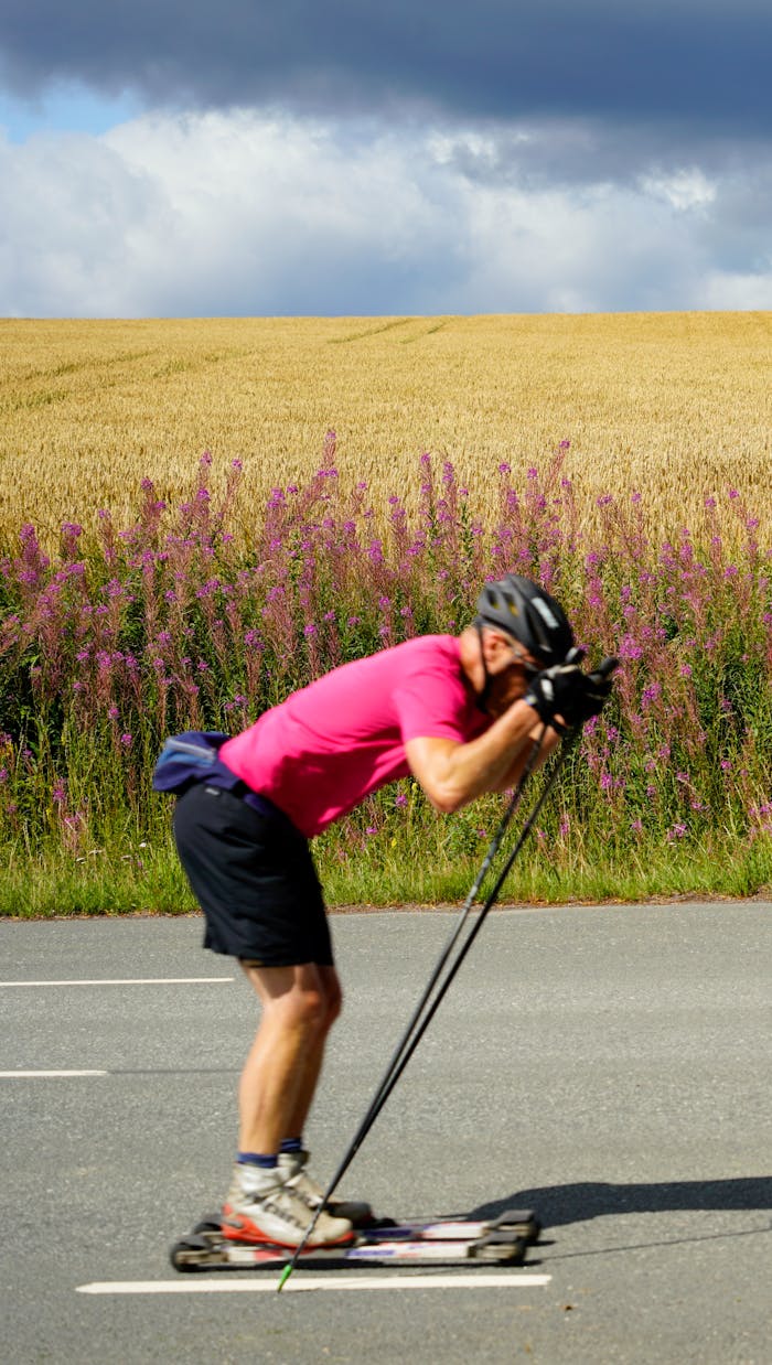 A man in a pink shirt roller skiing on a rural road with a field and flowers in the background.